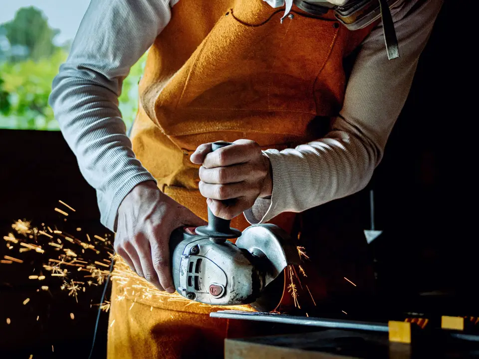 Un artisan en tablier de cuir découpe une pièce de métal à la meuleuse, dans une gerbe d’étincelles